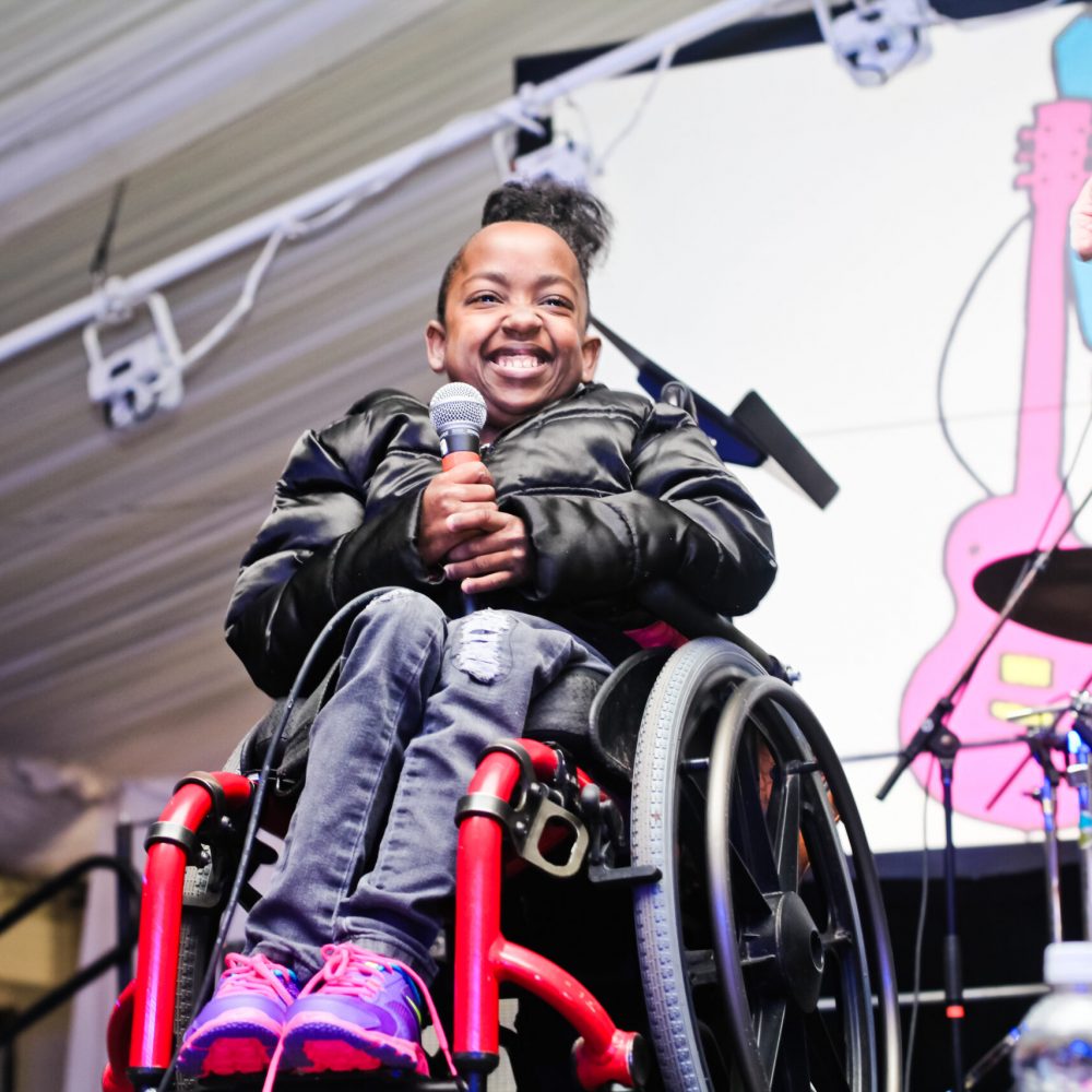A Black girl in a wheelchair is onstage, smiling and holding a microphone